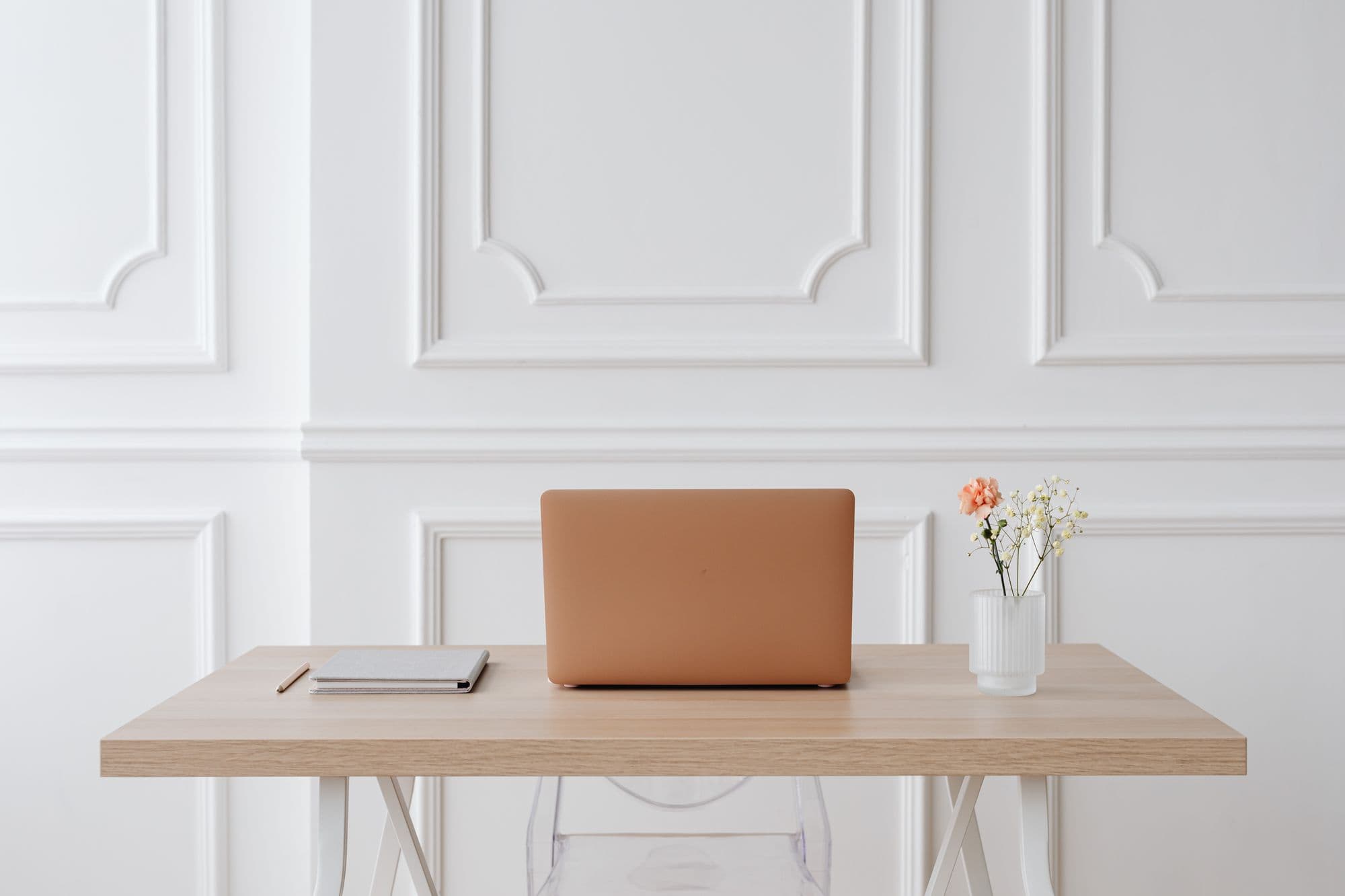 A minimal workspace with a copper laptop on a light-wood desk against white paneling